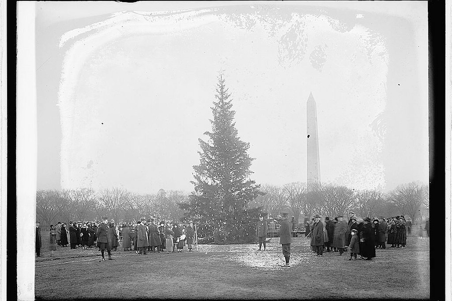 Black and white photo with people standing around a large Christmas tree. The Washington Monument is visible in the background.