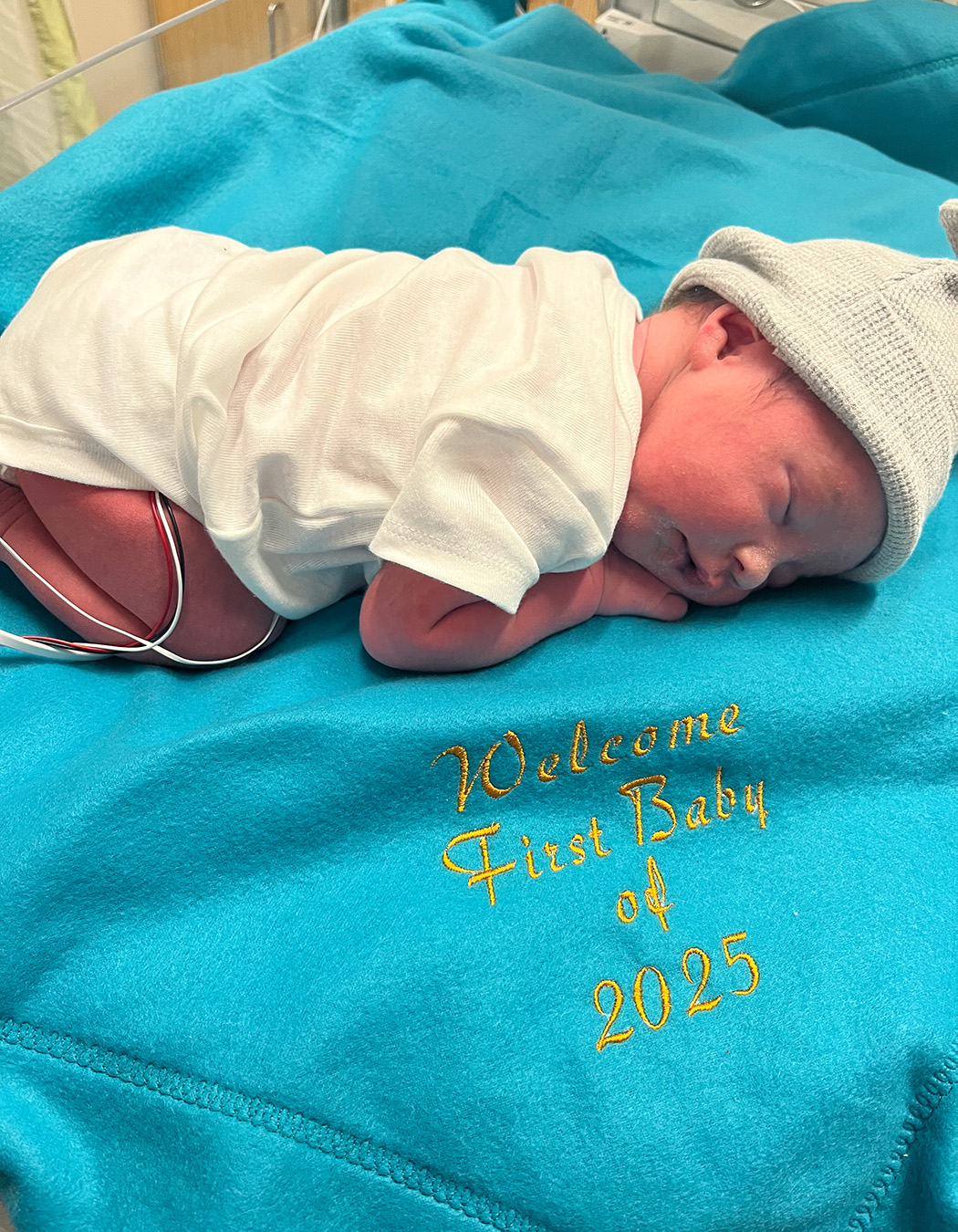 A newborn baby laying on a blanket with the sewn-in lettering that says, "Welcome first baby of 2025".