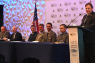 A man standing behind a podium that reads, "NPC Washington summit" with a board of individuals to his right.